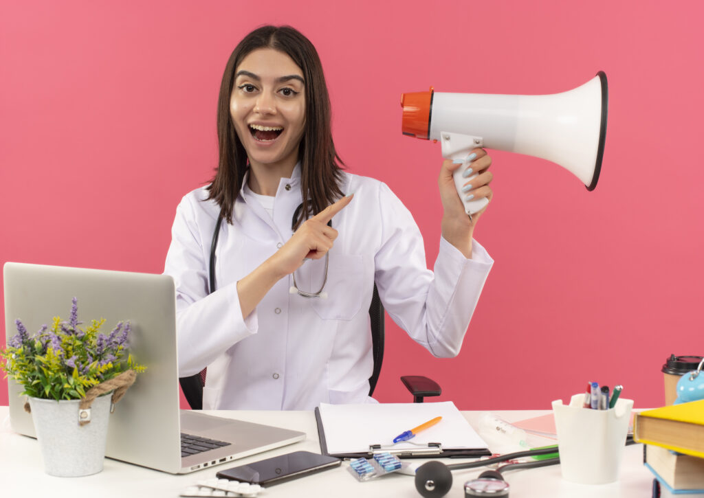 young-female-doctor-white-coat-with-stethoscope-around-her-neck-holding-megaphone-pointing-with-finger-it-smiling-cheerfully-sitting-table-with-laptop-pink-wall